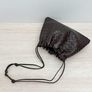 K&ouml;nnte beinhalten: Eine dunkelbraune Beuteltasche mit einem komplexen geflochtenen Ledermuster. Die Tasche hat eine geraffte &Ouml;ffnung im Kordelzug-Stil und einen d&uuml;nnen, verstellbaren Schulterriemen aus Leder mit einem kleinen Metalldruckknopf als Verschlussdetail.