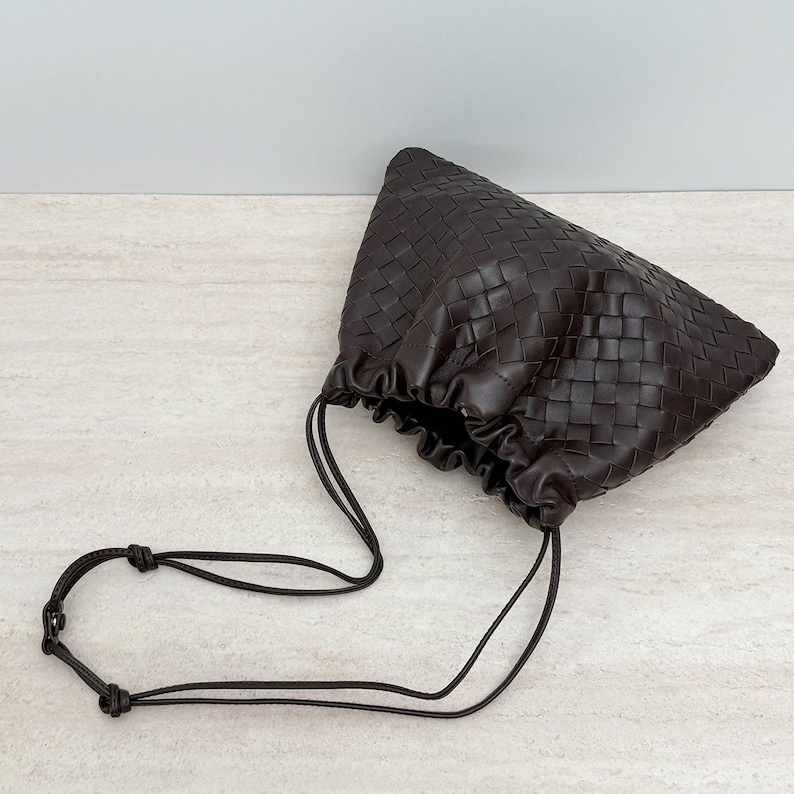 K&ouml;nnte beinhalten: Eine dunkelbraune Beuteltasche mit einem komplexen geflochtenen Ledermuster. Die Tasche hat eine geraffte &Ouml;ffnung im Kordelzug-Stil und einen d&uuml;nnen, verstellbaren Schulterriemen aus Leder mit einem kleinen Metalldruckknopf als Verschlussdetail.