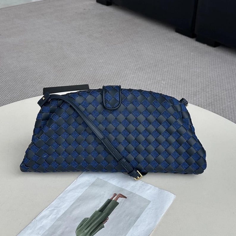 Op de afbeelding: Een blauw-zwarte geweven clutch tas met een schouderriem. De tas heeft een geometrisch patroon en een klepsluiting. De riem is verstelbaar met een goudkleurige gesp.
