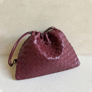 K&ouml;nnte beinhalten: Eine tiefburgunderrote Beuteltasche mit einem komplexen geflochtenen Ledermuster auf der gesamten Au&szlig;enseite. Die Tasche hat einen gerafften Kordelzugverschluss mit d&uuml;nnen Lederriemen, was eine weiche, lockere Silhouette mit einem strukturierten Boden erzeugt.
