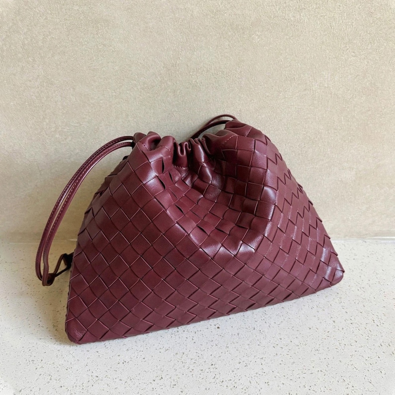 K&ouml;nnte beinhalten: Eine tiefburgunderrote Beuteltasche mit einem komplexen geflochtenen Ledermuster auf der gesamten Au&szlig;enseite. Die Tasche hat einen gerafften Kordelzugverschluss mit d&uuml;nnen Lederriemen, was eine weiche, lockere Silhouette mit einem strukturierten Boden erzeugt.
