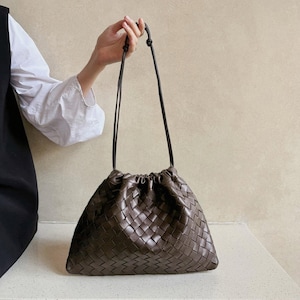 Könnte beinhalten: Eine dunkelbraune Handtasche mit geflochtenem Lederaußenmaterial und einem gerafften Kordelzugverschluss. Die Tasche verfügt über dünne, passende braune Lederschulterriemen, die oben verknotet sind und eine weiche, lockere Silhouette bilden.
