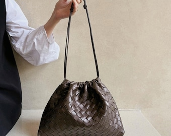 May include: A dark brown handbag featuring a woven leather exterior with a gathered drawstring top closure. The bag includes thin, matching brown leather shoulder straps that are knotted at the top, creating a soft, slouchy, and unstructured silhouette.