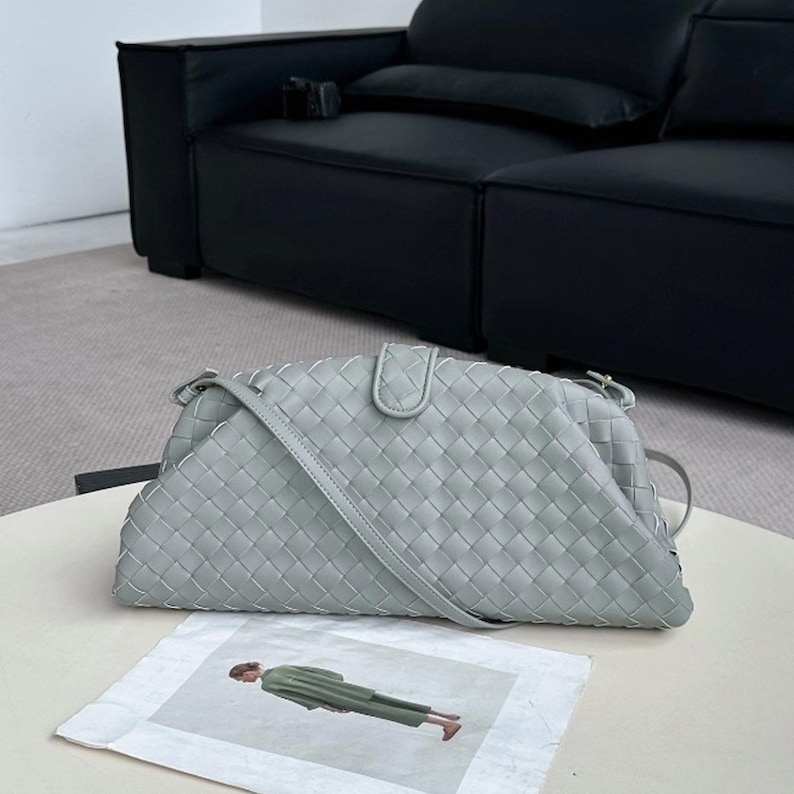 Op de afbeelding: Een lichtgroene geweven leren handtas met een schouderriem. De tas heeft een driehoekige vorm met een klepsluiting. De tas ligt op een witte tafel. Een zwarte bank staat op de achtergrond.