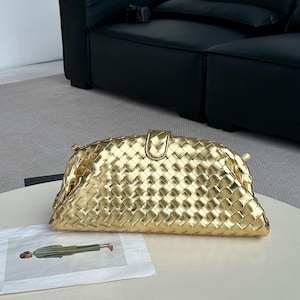 Op de afbeelding: Een goudkleurige clutch met een geweven ontwerp. De tas heeft een afgeronde vorm met een klepsluiting en een klein handvat. Hij ligt op een wit oppervlak.