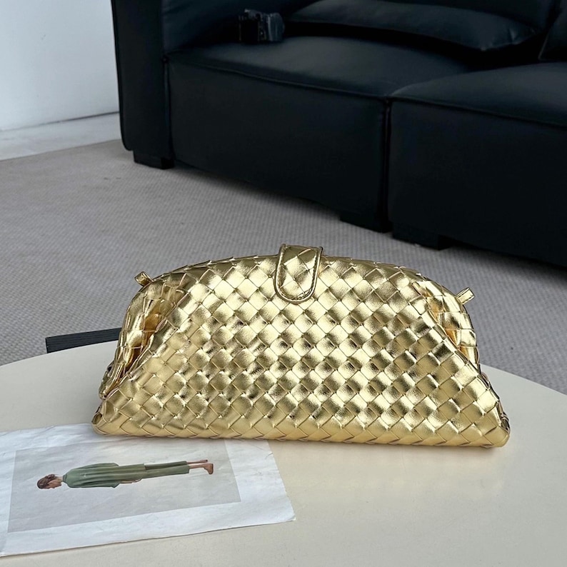 Op de afbeelding: Een goudkleurige clutch met een geweven ontwerp. De tas heeft een afgeronde vorm met een klepsluiting en een klein handvat. Hij ligt op een wit oppervlak.