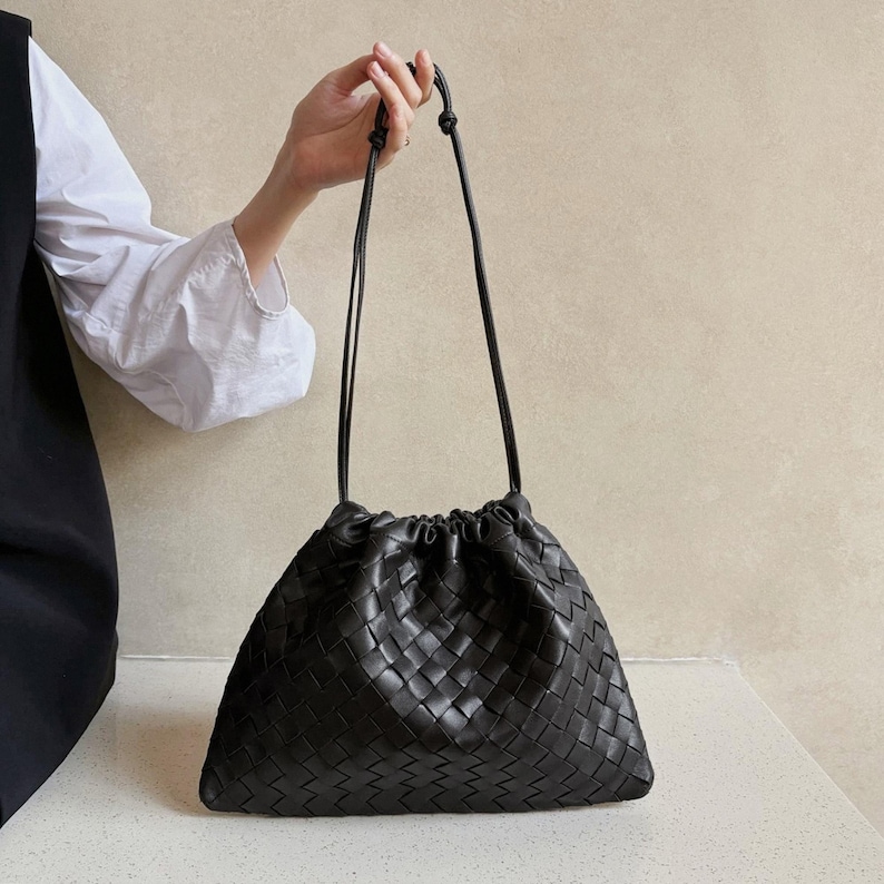 K&ouml;nnte beinhalten: Eine schwarze Handtasche mit geflochtenem Lederau&szlig;enmaterial, Kordelzugverschluss und d&uuml;nnen Schulterriemen. Die Tasche hat eine weiche, lockere Struktur und ein durchgehendes gitterartiges Muster aus ineinander verschlungenen Lederstreifen.