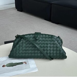 Op de afbeelding: Een donkergroene geweven clutch met een lange schouderriem. De clutch heeft een rechthoekige vorm met een gebogen bovenkant en een klepsluiting. Het geweven patroon bedekt het gehele oppervlak. De tas ligt op een witte tafel.