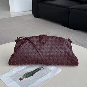 Op de afbeelding: Een bordeauxrode geweven leren clutch met een schouderriem. De tas heeft een rechthoekige vorm en een klepsluiting. De tas ligt op een wit oppervlak.