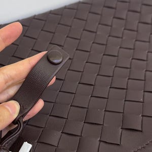 K&ouml;nnte beinhalten: Eine dunkelbraune Clutch mit einem geflochtenen Gittermuster auf der Oberfl&auml;che. Die Tasche verf&uuml;gt &uuml;ber eine passende dunkelbraune Handschlaufe, die mit einem runden Druckknopf befestigt ist. Das Material wirkt glatt und einheitlich.