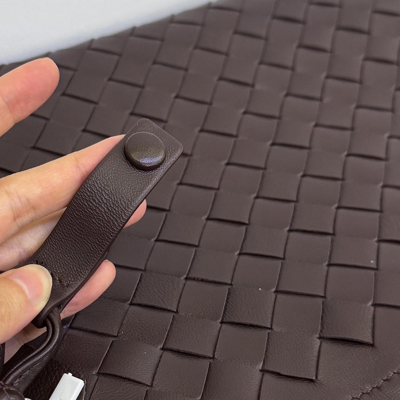 K&ouml;nnte beinhalten: Eine dunkelbraune Clutch mit einem geflochtenen Gittermuster auf der Oberfl&auml;che. Die Tasche verf&uuml;gt &uuml;ber eine passende dunkelbraune Handschlaufe, die mit einem runden Druckknopf befestigt ist. Das Material wirkt glatt und einheitlich.