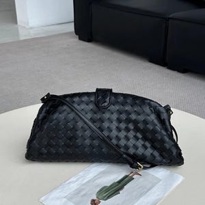Op de afbeelding: Zwarte geweven leren clutch met een lange schouderriem. De tas heeft een rechthoekige vorm met afgeronde randen en een klepsluiting. Het geweven patroon is zichtbaar over het gehele oppervlak. Een kleine goudkleurige gesp zet de riem vast.
