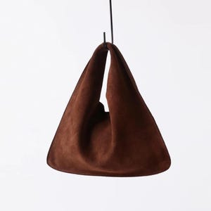 Puede incluir: Un bolso tote de ante marrón con forma triangular, suspendido de un gancho de metal negro. El bolso tiene un aspecto suave y texturizado y un diseño minimalista. Los bordes están cosidos. Altura aproximada: 40 cm.