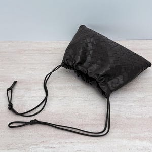 K&ouml;nnte beinhalten: Eine dunkelbraune Ledertasche mit einem komplexen Flechtmuster auf der Oberfl&auml;che. Die Tasche verf&uuml;gt &uuml;ber eine geraffte &Ouml;ffnung, die durch d&uuml;nne, passende Lederkordeln gesichert ist, welche nach au&szlig;en f&uuml;hren und in kleinen Knoten enden.