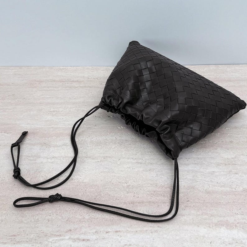 K&ouml;nnte beinhalten: Eine dunkelbraune Ledertasche mit einem komplexen Flechtmuster auf der Oberfl&auml;che. Die Tasche verf&uuml;gt &uuml;ber eine geraffte &Ouml;ffnung, die durch d&uuml;nne, passende Lederkordeln gesichert ist, welche nach au&szlig;en f&uuml;hren und in kleinen Knoten enden.