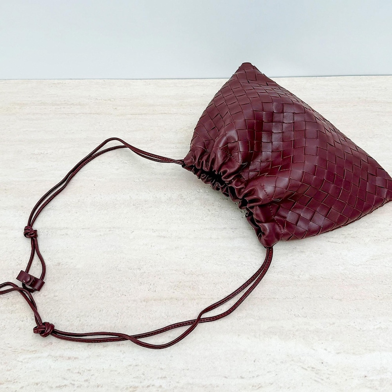 K&ouml;nnte beinhalten: Eine tiefburgunderrote Beuteltasche mit geflochtenem Lederau&szlig;enmaterial und geraubtem Verschluss. Die Tasche verf&uuml;gt &uuml;ber eine d&uuml;nne, passende burgunderrote Lederkordel, die nach au&szlig;en verl&auml;uft und mit Knoten sowie einem kleinen Schieberegler endet.
