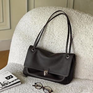 May include: A dark brown leather shoulder bag with a pebbled texture, featuring a fold-over flap, a silver-toned metal push-lock closure, and two thin shoulder straps attached to the bag with silver-toned metal rings.