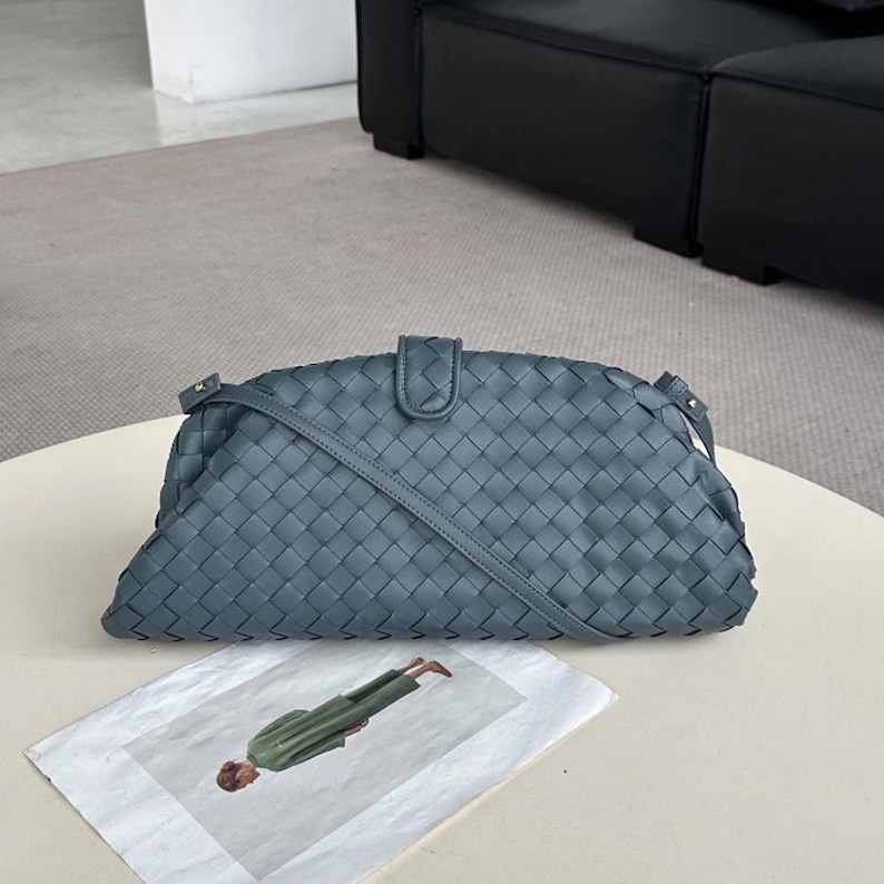 Op de afbeelding: Een geweven, leisteenblauwe handtas met een schouderriem. De tas heeft een rechthoekige vorm met een gebogen bovenkant en een klepsluiting. De tas staat op een witte tafel. Een tijdschrift met een persoon in groene kleding ligt op de tafel.