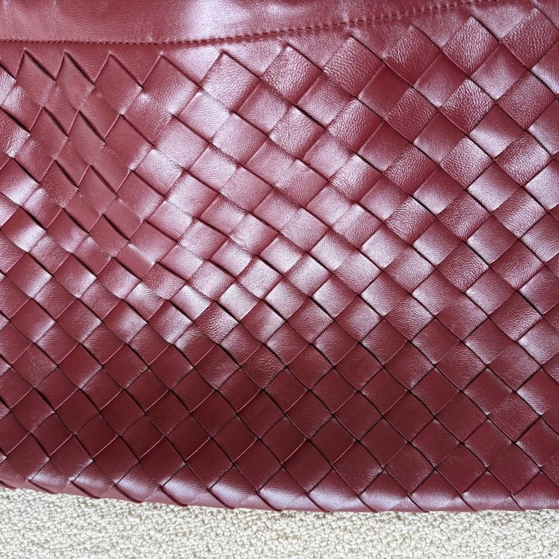 K&ouml;nnte beinhalten: Eine tiefburgunderrote Handtasche mit einer geflochtenen Lederkonstruktion in einem diagonalen Gittermuster. Die Oberfl&auml;che weist eine glatte, gleichm&auml;&szlig;ige Textur &uuml;ber die ineinandergreifenden Lederstreifen auf, abgeschlossen mit einer geraden Naht.