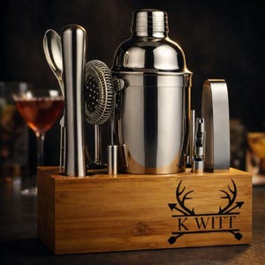 May include: A stainless steel cocktail shaker set with various bar tools, including a strainer, spoon, and jigger, presented in a wooden box. The box is engraved with a deer antler design and the name "K WITT".