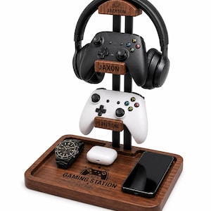 May include: A wooden gaming organizer stand featuring a base with the engraved text DAD'S GAMING STATION EST. 2026. The stand holds black headphones, a black controller labeled JAXON, and a white controller labeled PLAYER ONE. A watch, earbuds, and phone rest on the base.