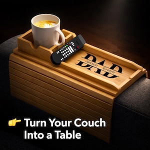 May include: A bamboo couch arm tray with a built-in cup holder and a remote control. The tray is engraved with the word "DAD" and the names "Jaxson" and "Evelyn". A white mug with coffee sits in the cup holder. The text "Turn Your Couch Into a Table" is at the bottom.