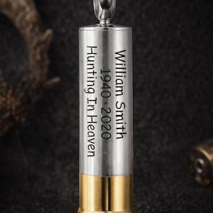 May include: A silver-colored bullet-shaped pendant necklace with a gold-colored base. The pendant has the engraved text "William Smith 1940-2020 Hunting In Heaven". The necklace is suspended from a silver chain.