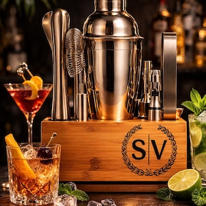 May include: A complete cocktail bar set with a stainless steel shaker, muddler, strainer, and jiggers, presented in a wooden box. The box is engraved with the letters "SV" inside a laurel wreath. Cocktails are visible in the foreground.