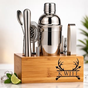 May include: A stainless steel cocktail shaker set with various bar tools, including a strainer, spoon, and tongs, presented in a wooden box. The box is engraved with a deer antler design and the name "K WITT".