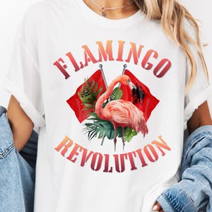 May include: A white t-shirt featuring a graphic of a pink flamingo standing in front of two crossed red flags and green tropical leaves. The text FLAMINGO REVOLUTION is printed in a red to orange gradient font above and below the central graphic.