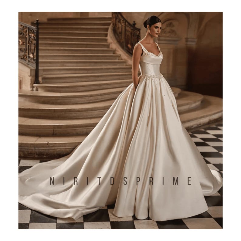 K&ouml;nnte beinhalten: Eine Frau in einem wei&szlig;en Satin-Hochzeitskleid mit einem herzf&ouml;rmigen Ausschnitt und einem vollen Rock. Das Kleid hat einen G&uuml;rtel mit Perlen und einen Schleier. Die Frau steht auf einer Treppe mit einem karierten Boden.