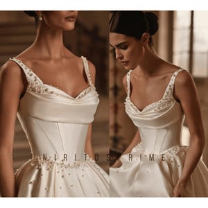 K&ouml;nnte beinhalten: Ein wei&szlig;es Satin-Hochzeitskleid mit einem herzf&ouml;rmigen Ausschnitt und einem taillierten Oberteil. Das Kleid hat einen vollen Rock und ist mit Perlen verziert.