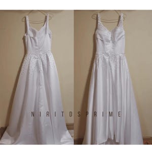 K&ouml;nnte beinhalten: Ein wei&szlig;es Satin-Hochzeitskleid mit einem herzf&ouml;rmigen Ausschnitt und einem perlenbesetzten Oberteil. Das Kleid hat einen hohen, niedrigen Saum, wobei der vordere Saum &uuml;ber dem Knie endet und der hintere Saum bis zum Boden reicht.