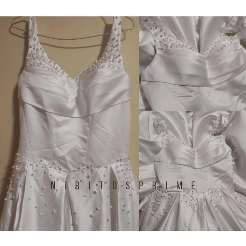 K&ouml;nnte beinhalten: Ein wei&szlig;es Satin-Hochzeitskleid mit einem herzf&ouml;rmigen Ausschnitt und perlenbesetzten Tr&auml;gern. Das Kleid hat einen vollen Rock und ist mit Perlen verziert.