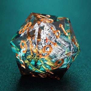 May include: A multi-faceted, clear resin die with gold-colored numbers and internal details. The die has a complex, layered design with teal and silver accents. A unique gaming accessory.