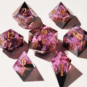 Könnte beinhalten: Sieben polyedrische Würfel für Tabletop-Spiele. Die Würfel bestehen aus einer Mischung aus schwarzem und durchscheinendem Harz mit wirbelnden rosa und weißen Mustern. Goldfarbene Zahlen sind auf jedem Würfel sichtbar.
