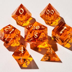 May include: Set of seven orange-colored polyhedral dice with white numbering. The dice are transparent and cast shadows on a white surface. The set includes a variety of shapes, including a d20, d12, d10, d8, d6, d4, and a d00.