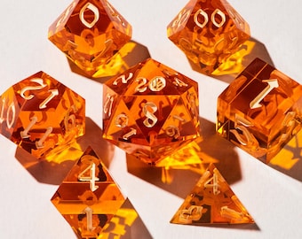 May include: Set of seven orange-colored polyhedral dice with white numbering. The dice are transparent and cast shadows on a white surface. The set includes a variety of shapes, including a d20, d12, d10, d8, d6, d4, and a d00.