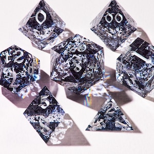 May include: Set of seven polyhedral dice for tabletop games. The dice are clear with a dark blue and silver speckled design. Each die has silver numbering. The dice are arranged on a white surface, casting shadows.