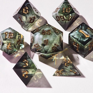 May include: Set of seven polyhedral dice for tabletop games. The dice are a dark green with white marbling and gold-colored numbers. The dice are arranged on a white surface, with shadows cast from the light source.
