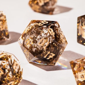 Może przedstawiać: Zbliżenie na kilka wielościennych kostek. Kości są przezroczyste z brązowo-złotym wnętrzem i złotymi numerami. Kości są rozrzucone na białej powierzchni, z jedną kostką w centrum, wyświetlającą numer 17.