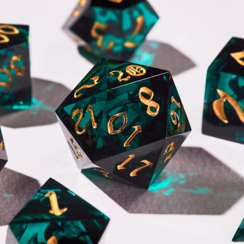 May include: Close-up of several polyhedral dice. The dice are emerald green with gold-coloured numbers. The dice are scattered on a white surface, with shadows cast by the light source. The dice are used for tabletop games.