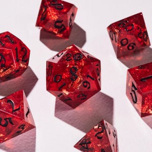 May include: A set of red polyhedral dice with black numerals and gold leaf inclusions. The dice are made from a transparent material, and the numbers are easily seen. The dice are displayed on a white surface.