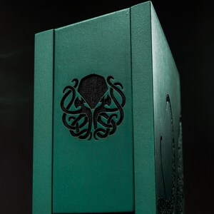 May include: A teal-colored wooden box with a carved Cthulhu design. The box features a circular cut-out with a black interior and a detailed tentacle design. The box is rectangular and has a textured surface.