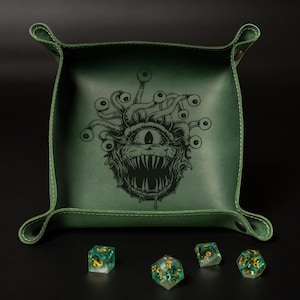 May include: A green leather dice tray with a detailed illustration of a monster with multiple eyes. The tray has a stitched border and rolled corners. Several multi-sided dice with gold accents are scattered around the tray.