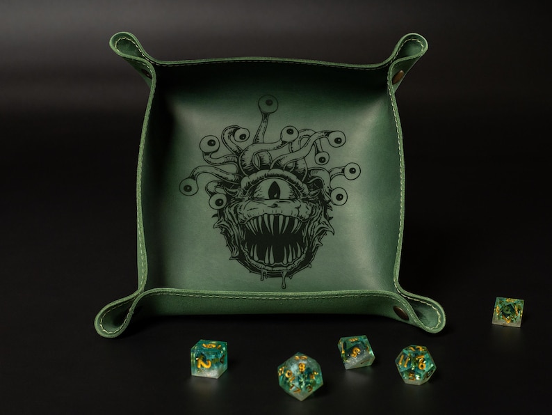 May include: A green leather dice tray with a detailed illustration of a monster with multiple eyes. The tray has a stitched border and rolled corners. Several multi-sided dice with gold accents are scattered around the tray.