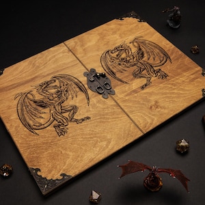 K&ouml;nnte beinhalten: Hellbraune Holzspielbox mit eingravierten Drachendesigns. Die Box hat dunkle Metallecken und einen Verschluss. W&uuml;rfel, Figuren und andere Spielsteine sind darum herum angeordnet, was auf ein Tabletop-Gaming-Thema hindeutet.