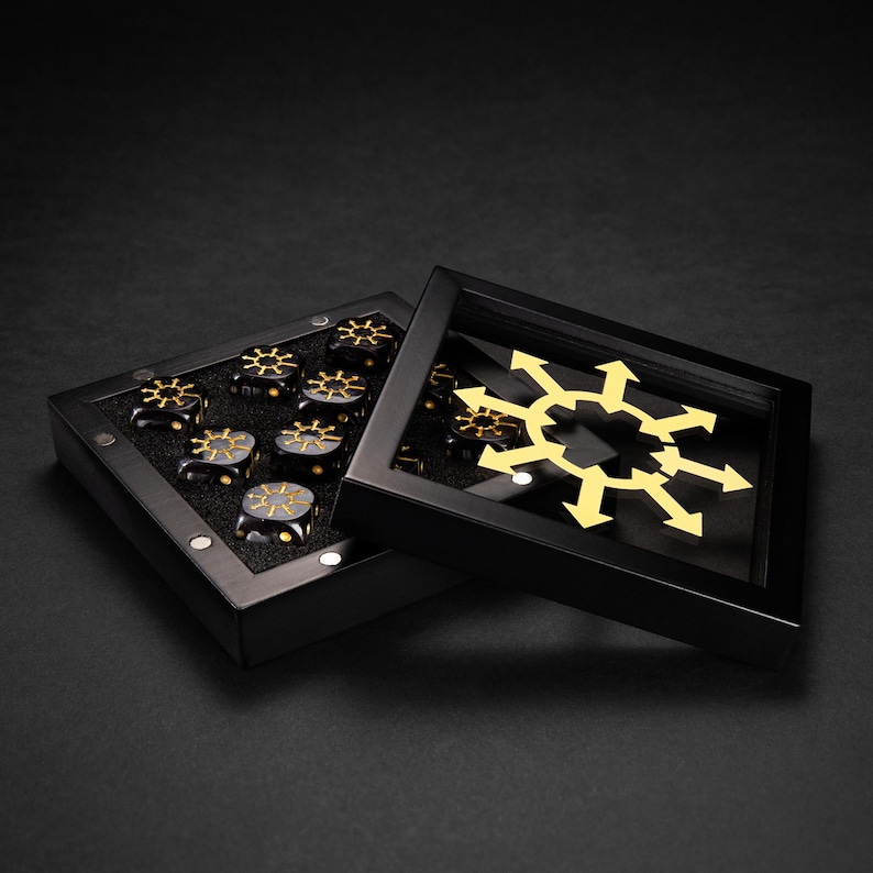 May include: A black box with a clear lid, containing several black dice with gold-colored symbols. The box is open, revealing the dice and a gold-colored symbol on the lid. The dice are arranged in a neat row.
