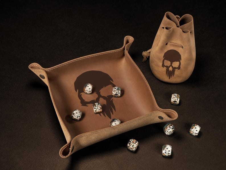 May include: A brown leather dice tray and matching drawstring bag, both featuring a skull design. Several speckled dice are scattered around the tray. The set is designed for tabletop gaming.