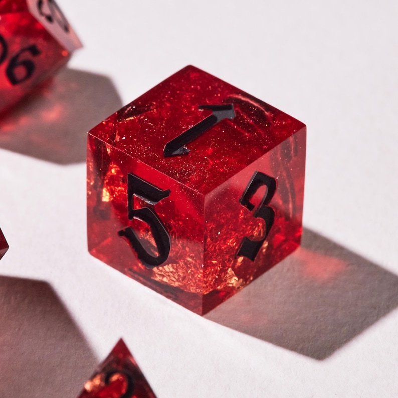May include: A close-up of a red, six-sided die with black numbers and a gold flecked interior. The die shows the numbers 5, 3, and 1. The die is made of a clear resin material.